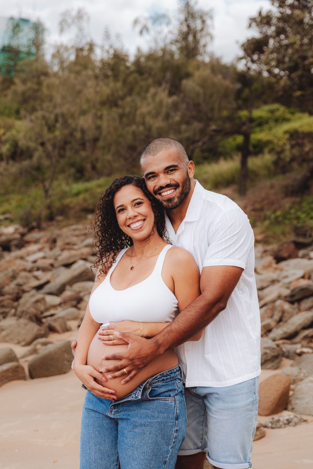 Happy pregnant couple embracing outdoors, woman in white crop top holding baby bump, man in white shirt smiling, rocky nature background.
