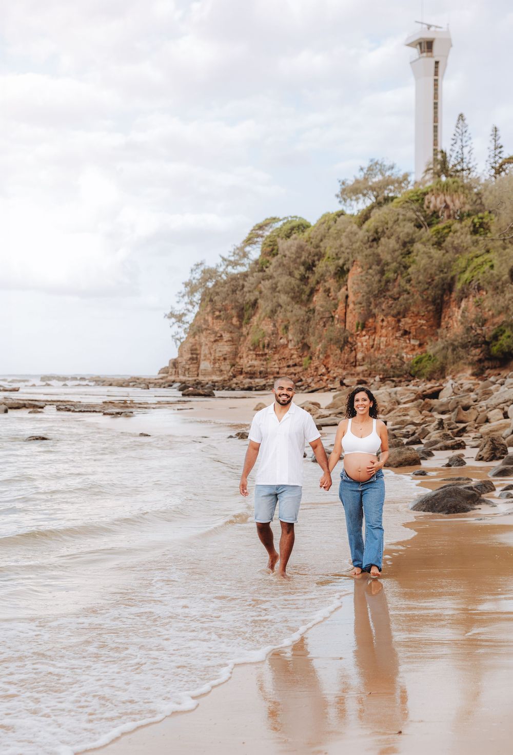 Pregnant couple walking hand in hand on a sandy beach with a lighthouse and rocky cliffs in the background.
