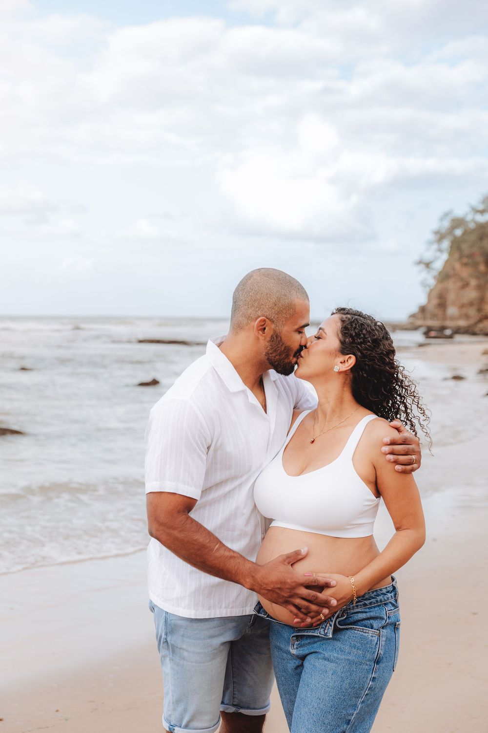 Pregnant couple kissing on a beach, man holding woman's baby bump, both wearing white tops and jeans.
