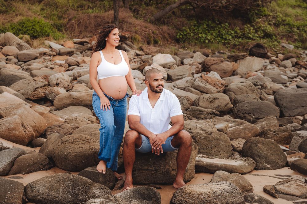 Pregnant couple posing on rocky beach, woman in white crop top and jeans, man in white shirt sitting on stone.