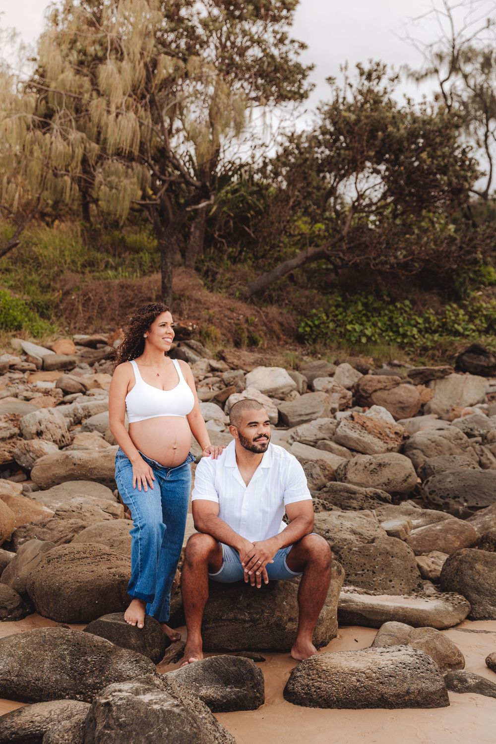 Pregnant couple posing on rocky beach, woman in white crop top and blue jeans, man seated in white shirt and blue shorts.