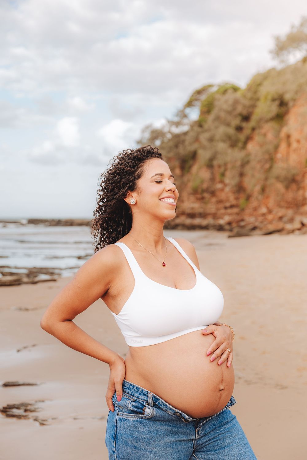Smiling pregnant woman in white crop top and jeans cradling her baby bump on a sandy beach with rocky cliffs.