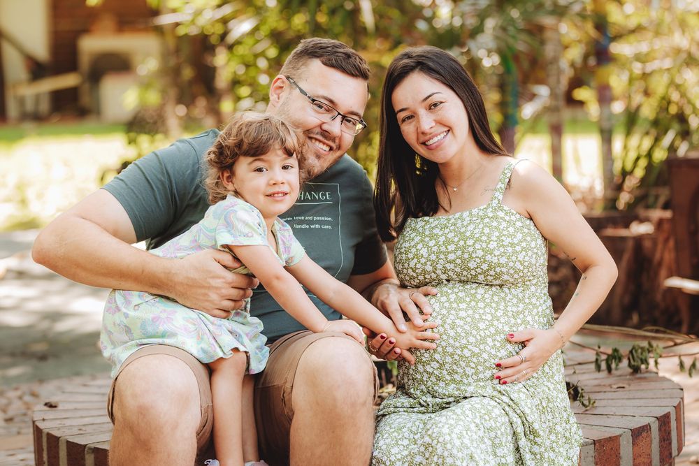 Smiling pregnant woman in green floral dress sits with husband and toddler daughter outdoors in sunny park setting.