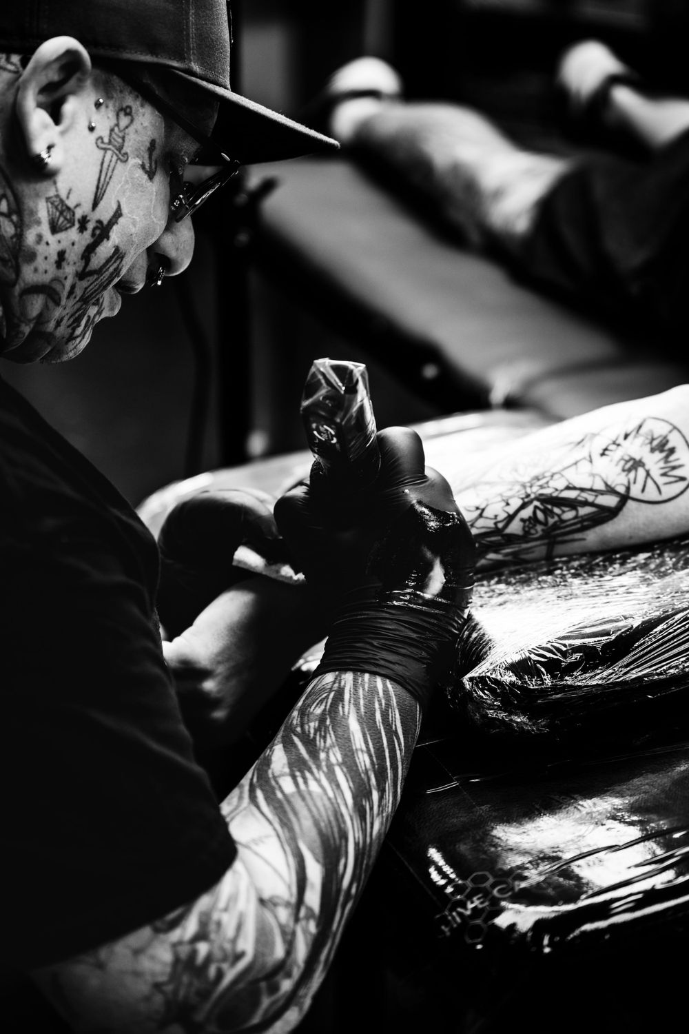 Black and white photograph of a tattoo artist at work using a tattoo machine, with plastic-wrapped equipment visible.