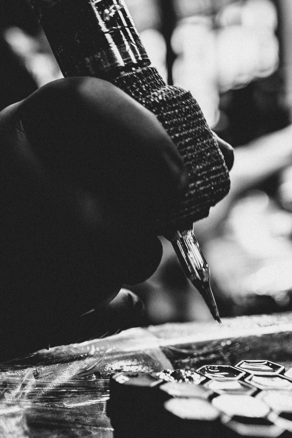 A black and white macro shot shows an artist's hand gripping a tattoo machine while working on a design.