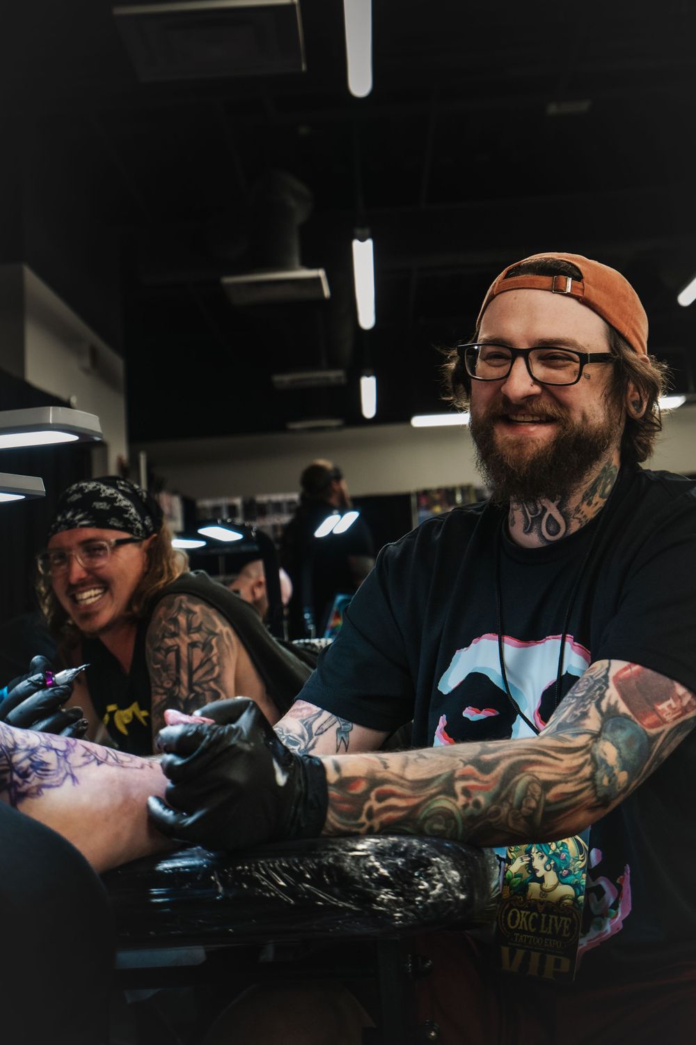 Tattoo artists wearing dark clothing and baseball caps work at a convention with their equipment and gloves.