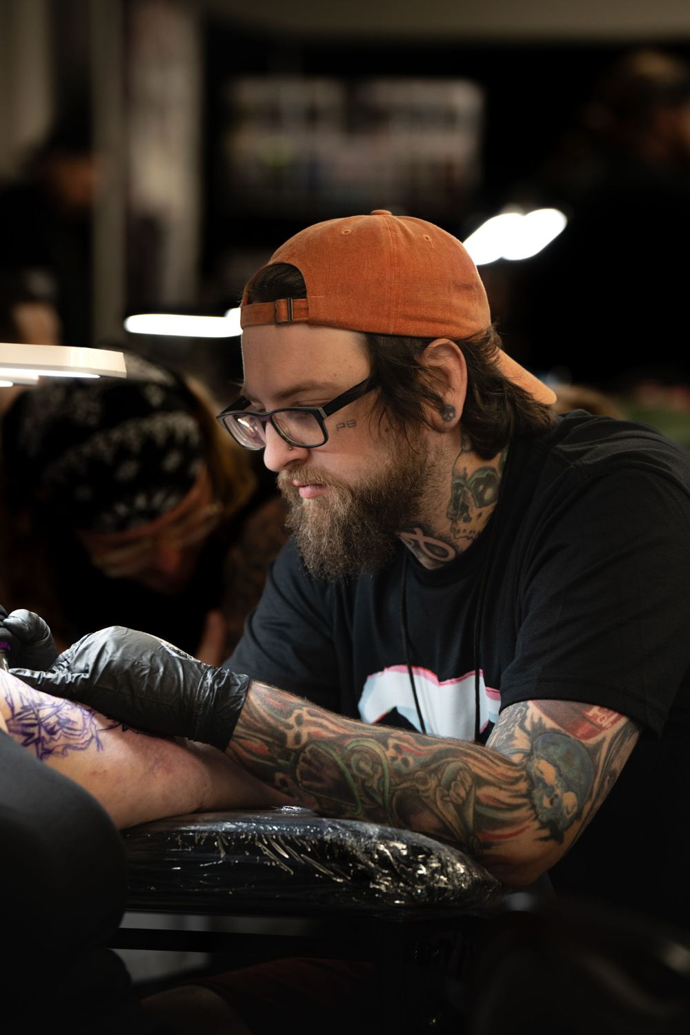 Professional tattoo artist with beard and orange cap works intently on a client's arm in a studio setting.