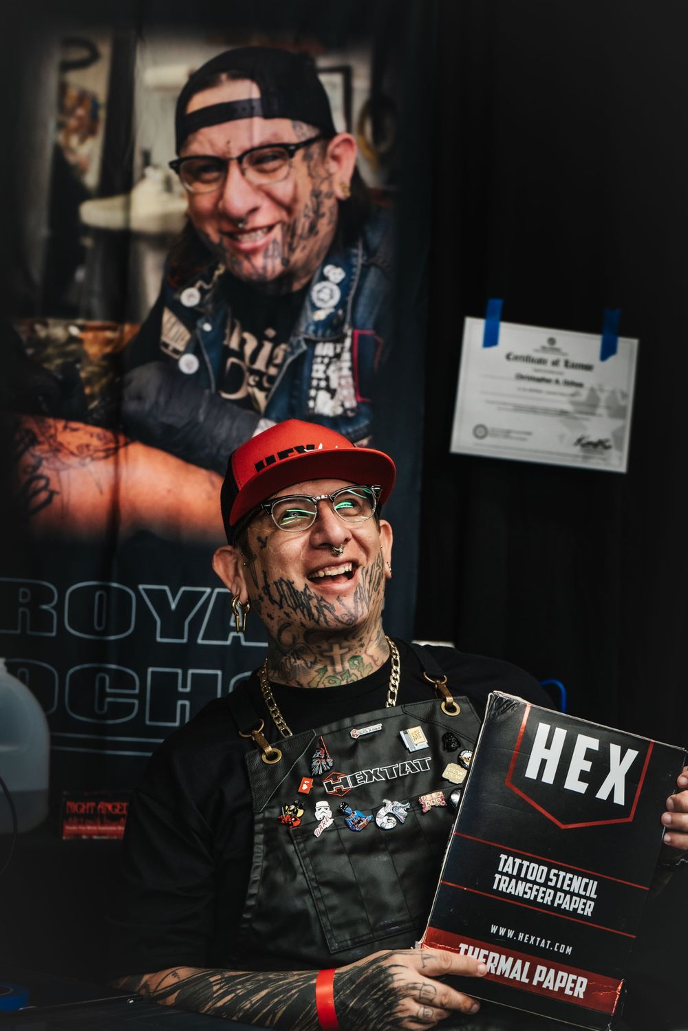 Tattooed artist wearing glasses and baseball cap smiles while holding a HEX brand tattoo stencil paper.
