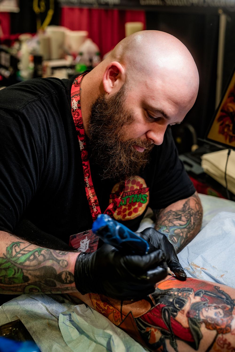 Professional tattoo artist with a full beard wearing black shirt and gloves works on a detailed tattoo.