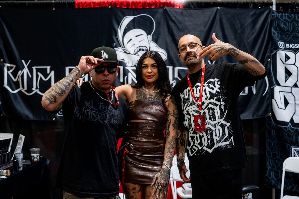 Three people pose together at a tattoo convention in front of a dark backdrop with event branding.