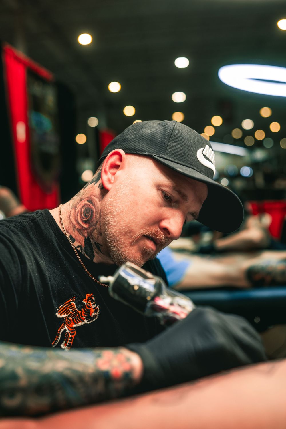 A tattoo artist wearing a black cap and t-shirt works intently on a client in a studio with bokeh lighting in the background.