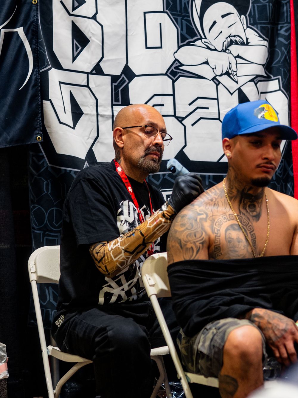 Tattoo artist works on a shirtless client with extensive body art against a graffiti wall backdrop at a tattoo convention.