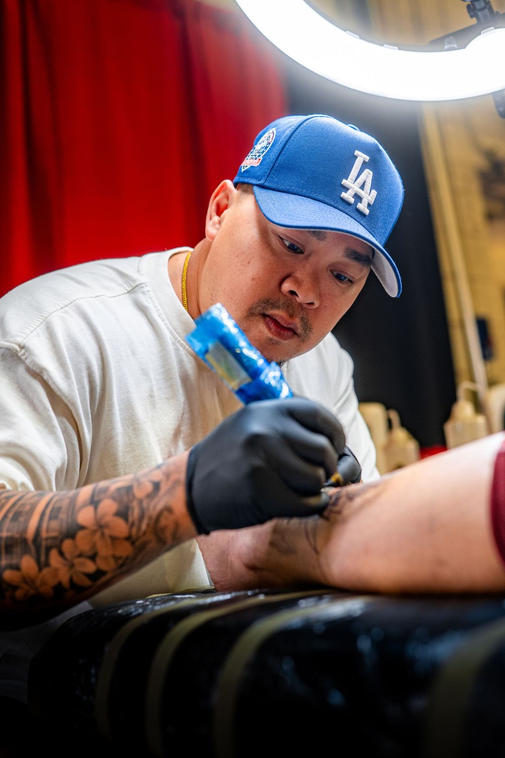 A tattoo artist wearing a blue LA Dodgers cap and black gloves works on a client in a professional studio setting.