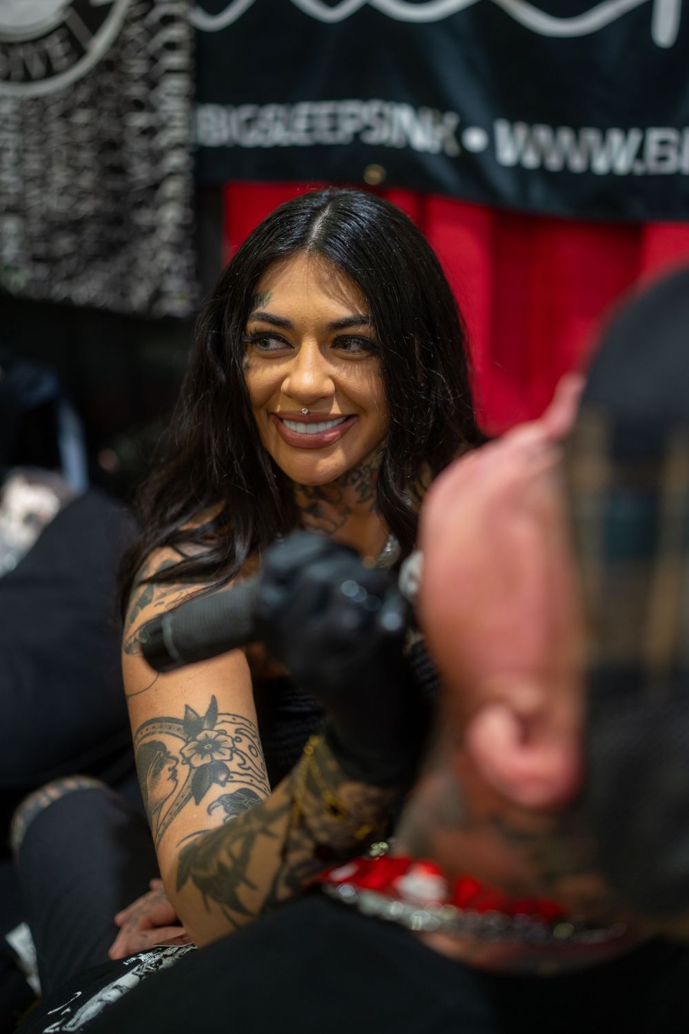 Portrait of a smiling person with long dark hair and visible arm tattoos in a dimly lit setting.