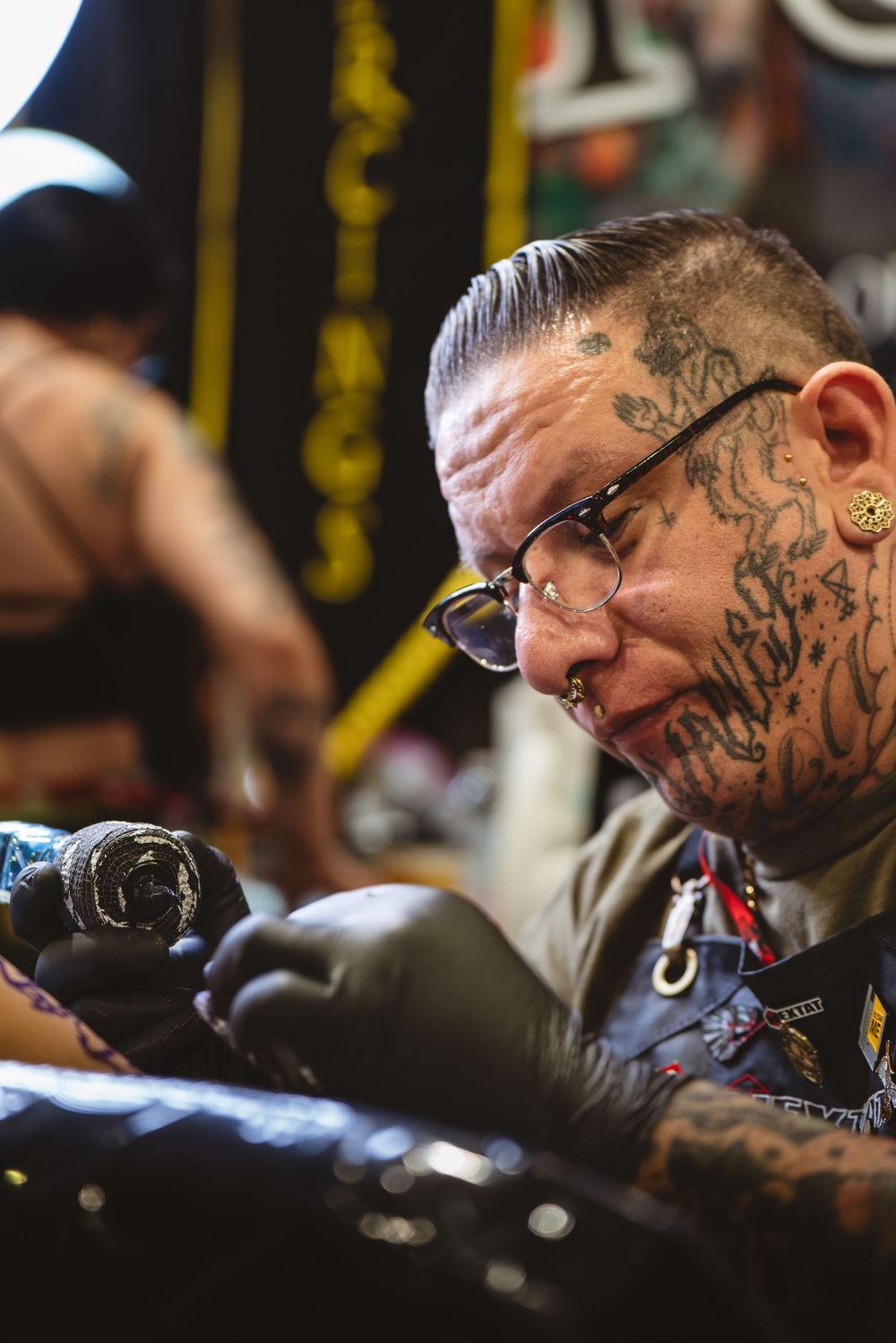 Tattoo artist wearing glasses concentrates intently while working on a client in a dimly lit studio setting.