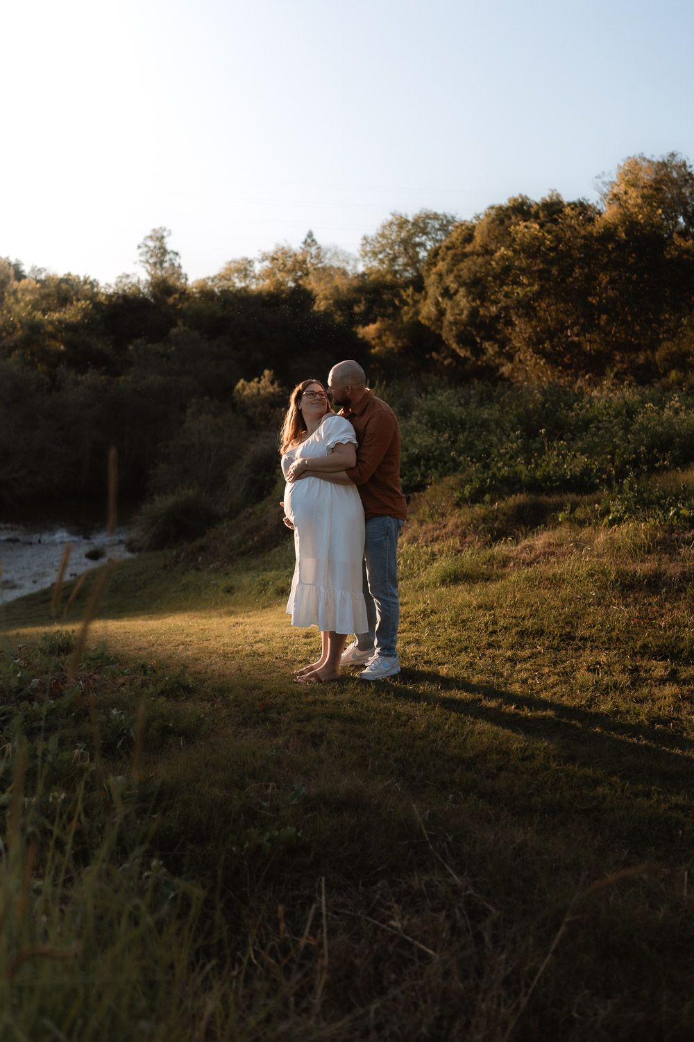 A couple embraces on a grassy hillside during golden hour as sunlight filters through trees in the background.