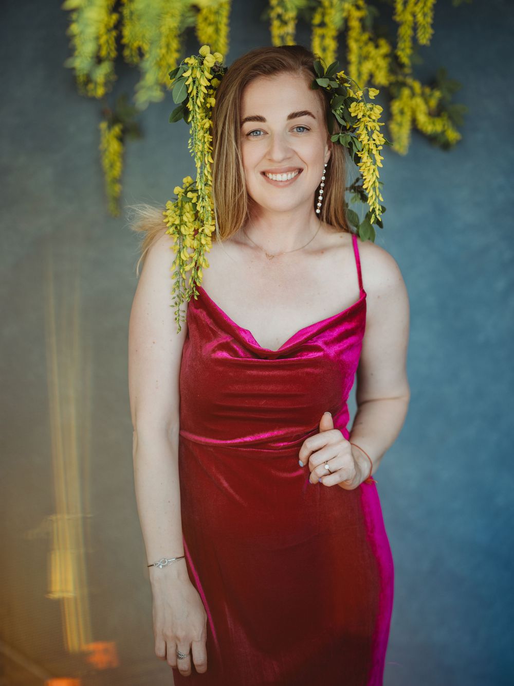 A person in a burgundy velvet dress poses under hanging yellow flowers against a blue backdrop.