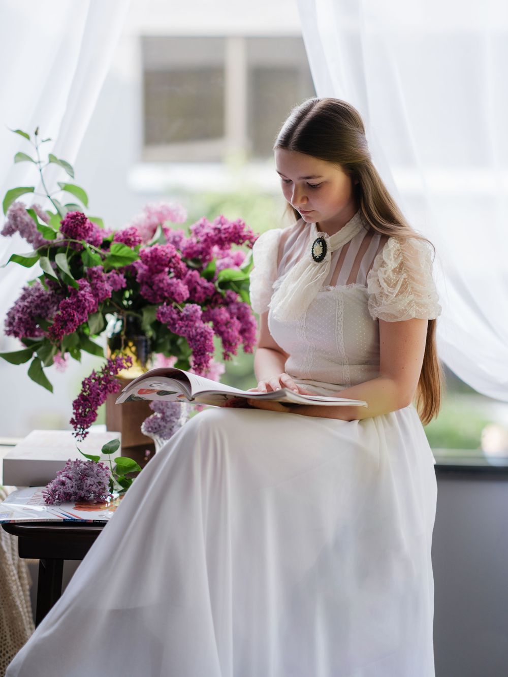A person in a white dress reads near a window with purple lilac flowers in an elegant indoor setting.