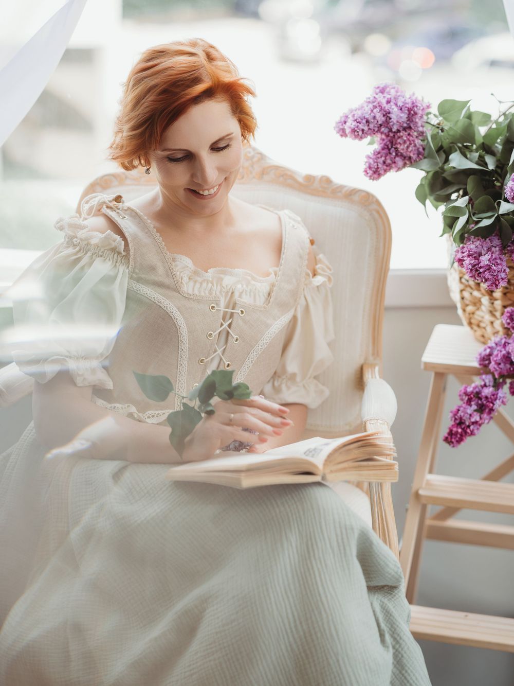 Woman in vintage-style white dress reads book near window with lilac flowers in elegant spring setting.