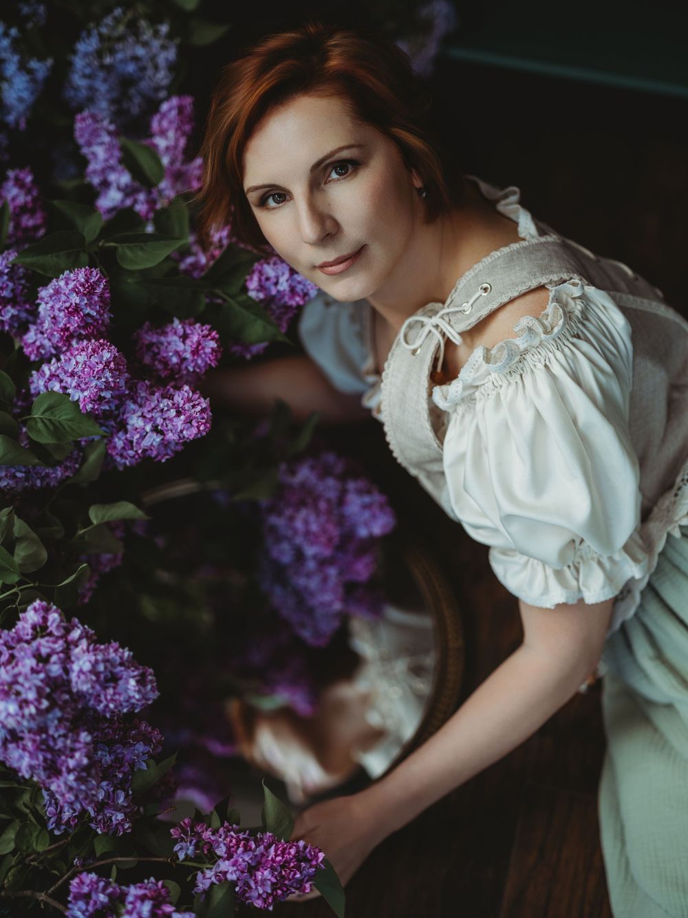 A person in a vintage white blouse poses gracefully against a backdrop of blooming purple lilac flowers in a garden setting.