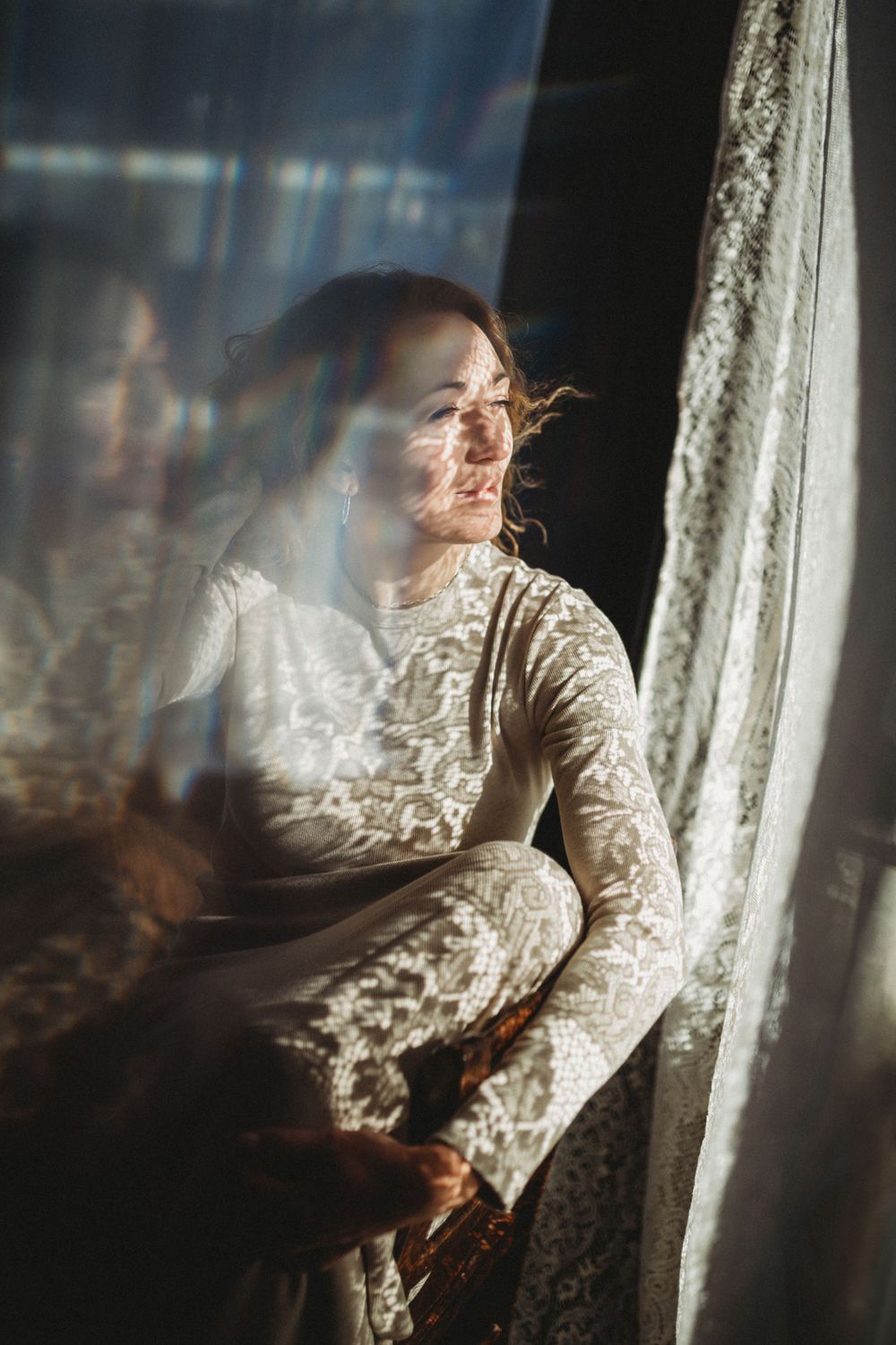 Soft light streams through lace curtains illuminating a person in a vintage dress by a window. Bellevue studio, WA