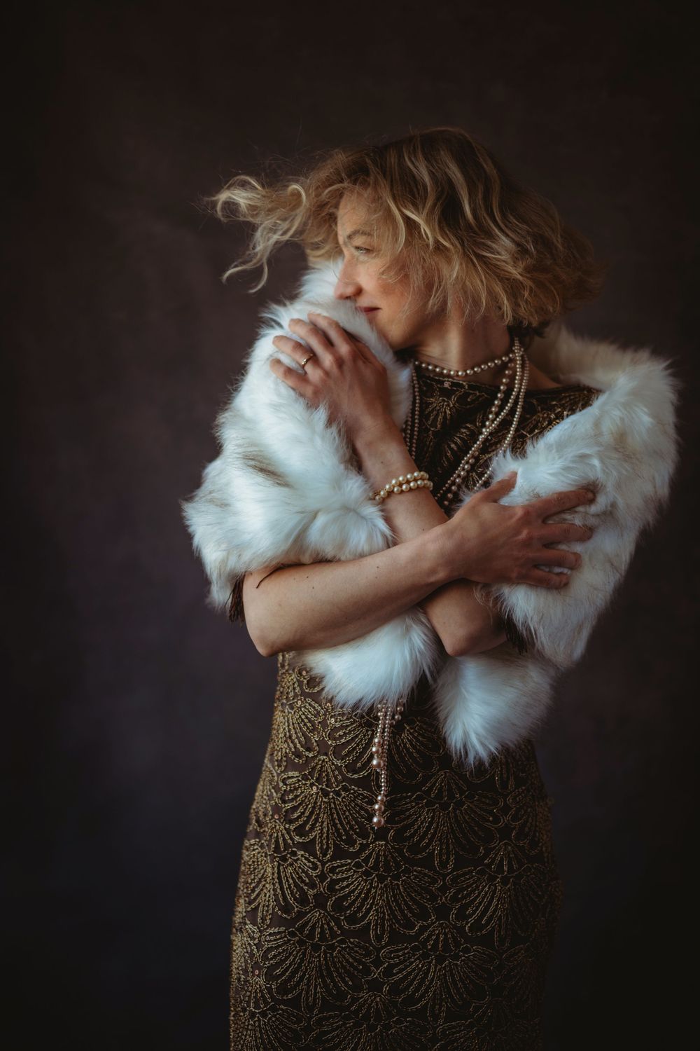 A figure in a vintage-style gold dress and white fur stole poses dramatically against a dark background in a photo series.