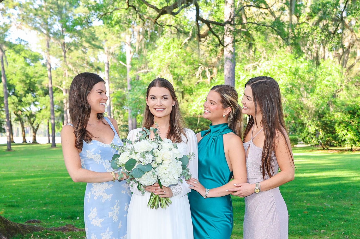 A bridal party in pastel dresses poses together on a green lawn under sunlit trees.