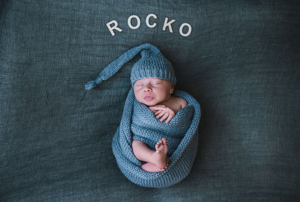 A newborn baby wearing a blue knit wrap lies curled up in different sleeping poses against a dark backdrop.