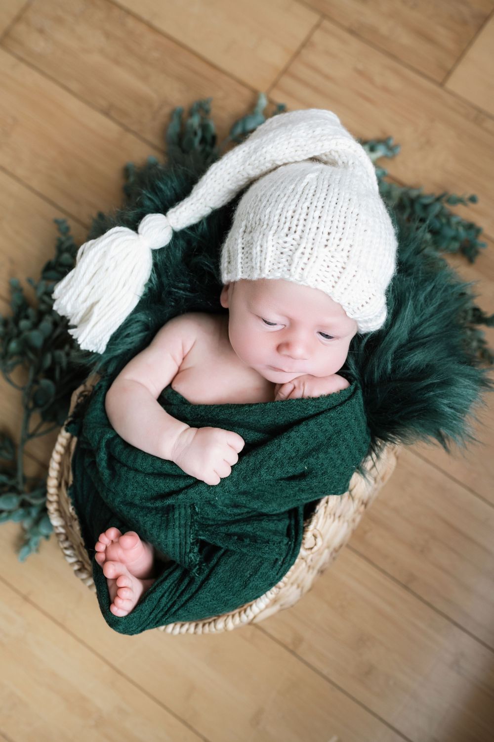 Newborn sleeping in basket with white knit hat and green wrap on wooden floor.