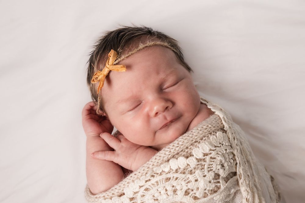Sleeping newborn baby wrapped in cream knit blanket wearing yellow floral headband.