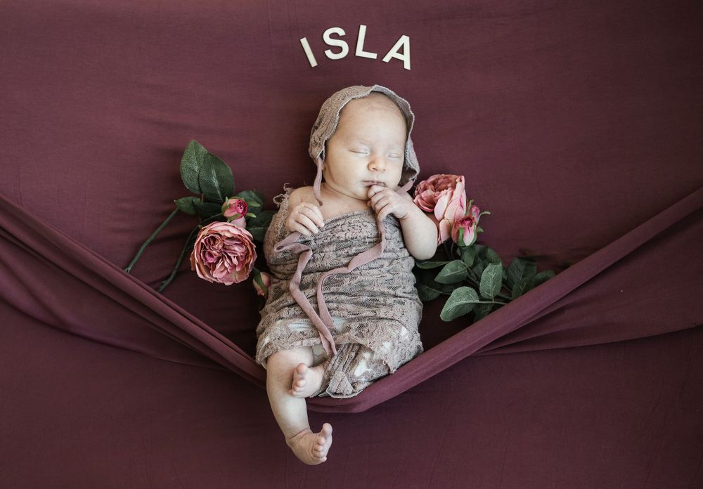 A newborn baby sleeps peacefully on burgundy fabric with pink roses and greenery decorations.