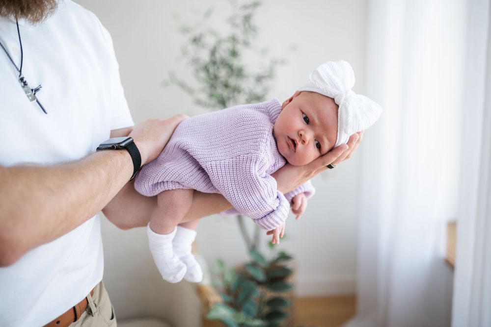Father holding newborn baby in white bow hat and pink knit outfit in bright nursery room.