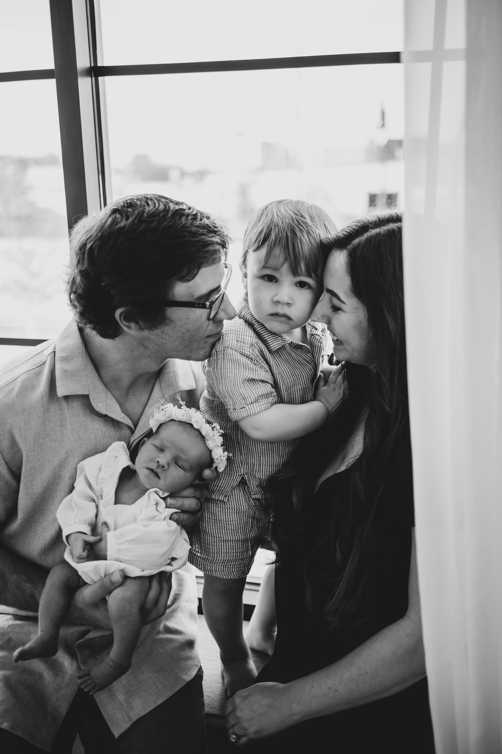 Black and white family portrait with parents kissing toddler while father holds sleeping newborn baby by window.