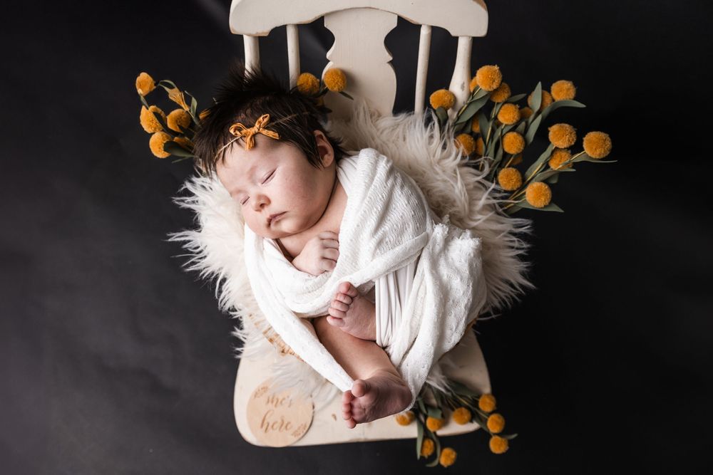 Sleeping newborn in white wrap on chair with orange flowers against dark background.