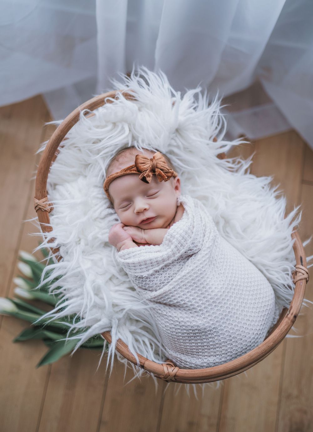 Newborn baby wrapped in white knit blanket sleeping in basket with tulips.