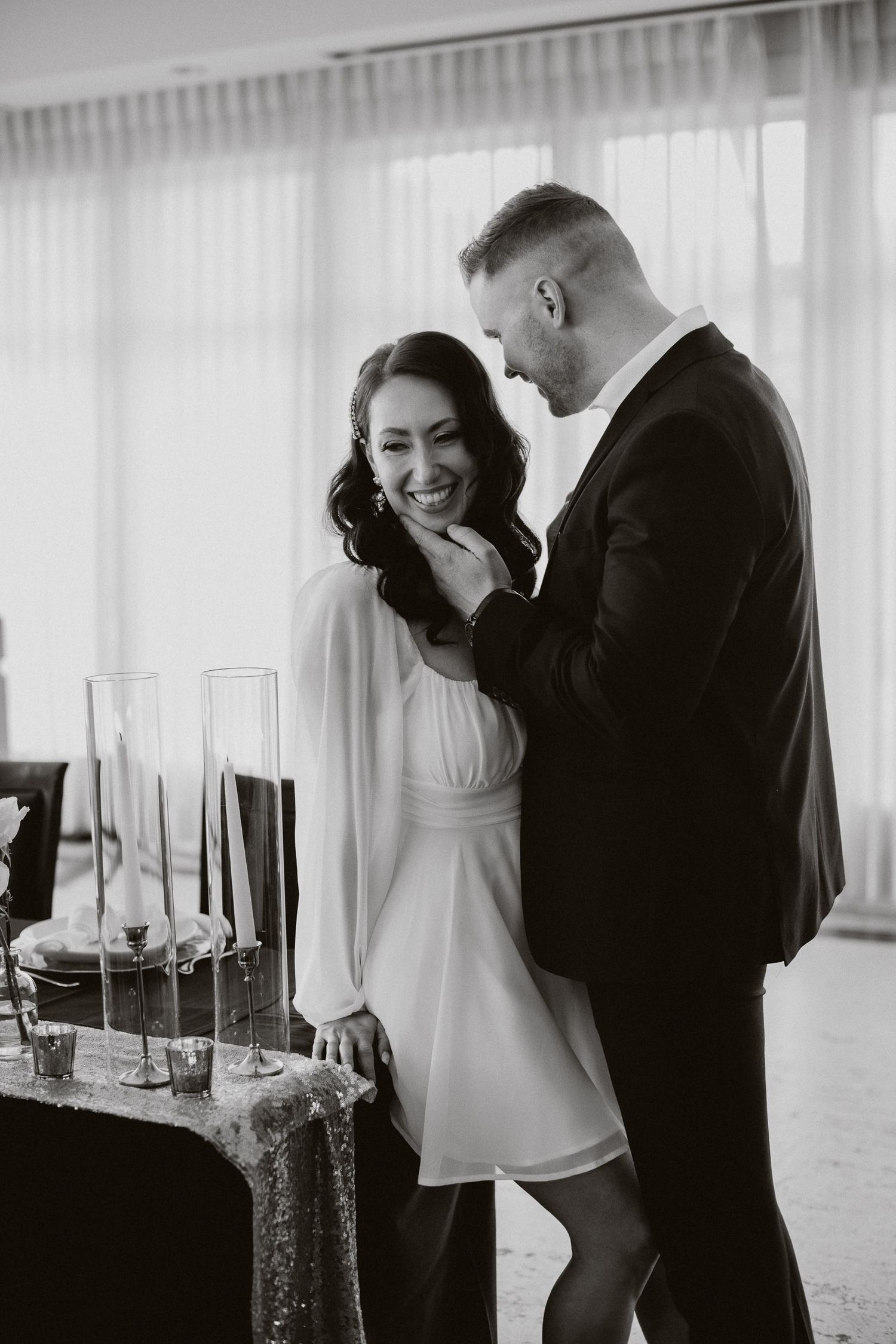 A couple shares a tender moment in black and white while standing next to a table with tall candlesticks.