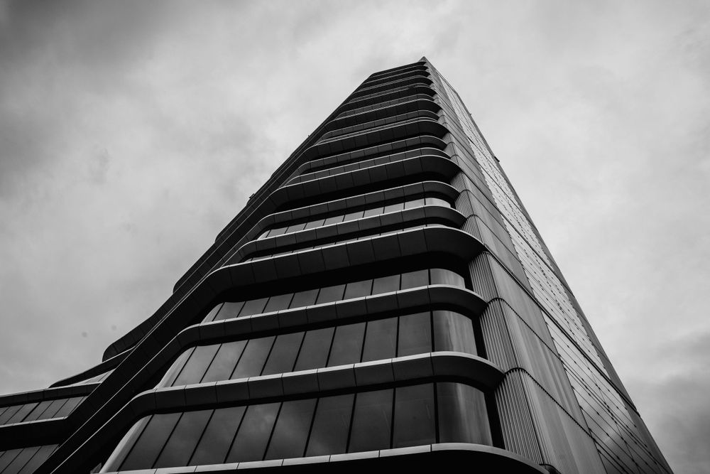 Modern glass skyscraper photographed from below in black and white against moody clouds.