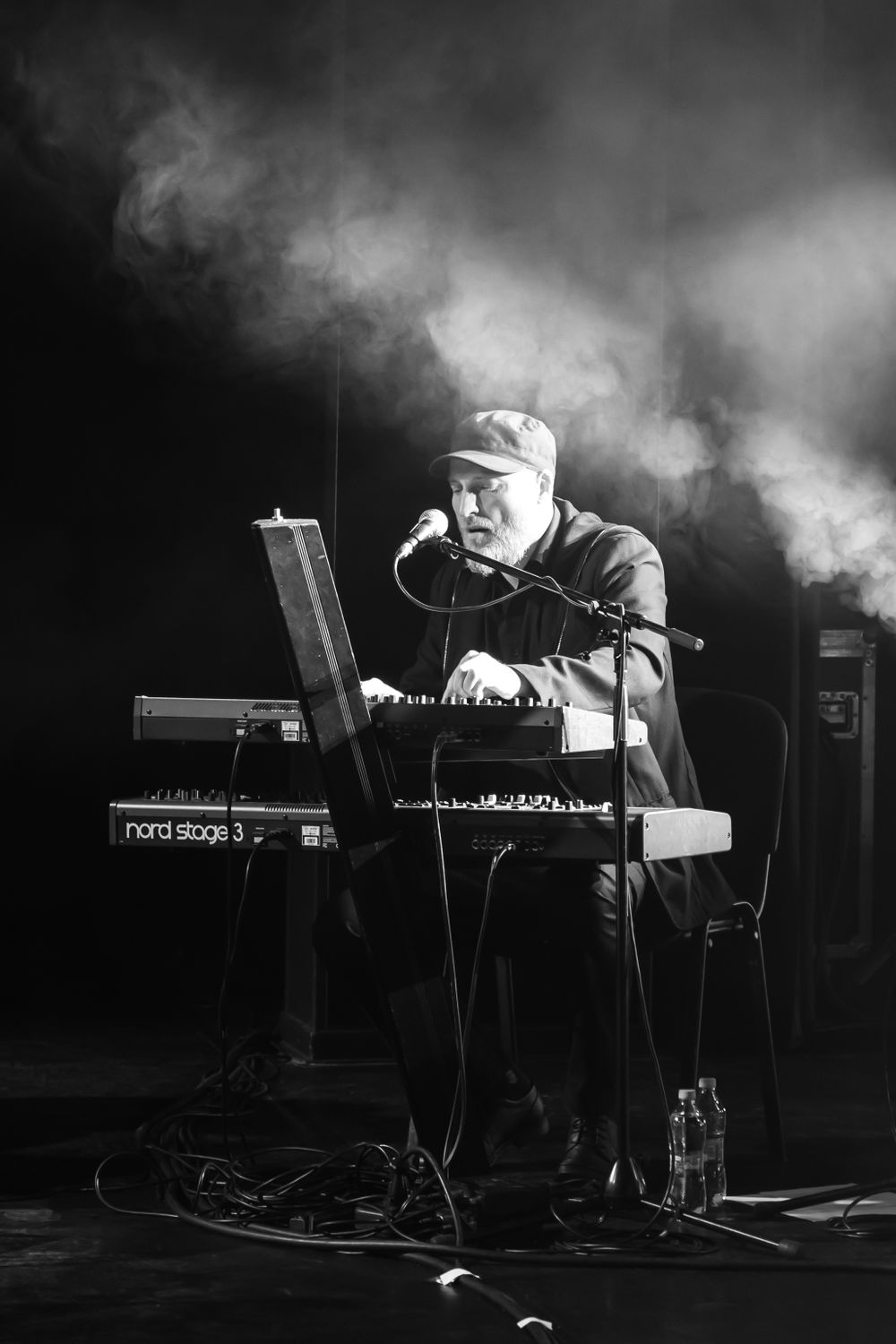 Musician performing with electronic equipment on a smoke-filled stage in dramatic black and white.