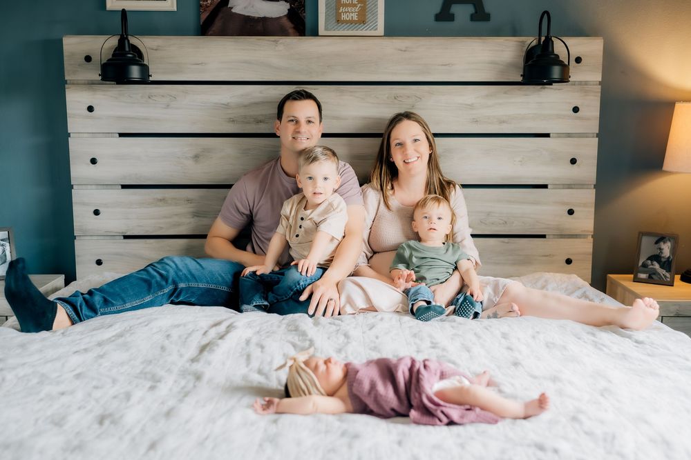 A family relaxes together on a white bed with a rustic wooden headboard in a cozy bedroom setting.