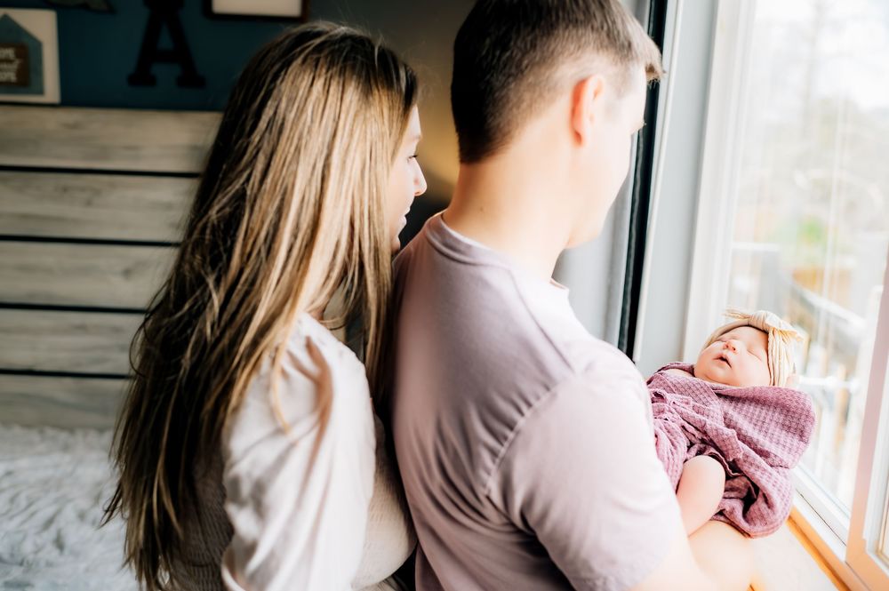 A couple stands by a window with their baby, looking out at natural light filtering through the glass.