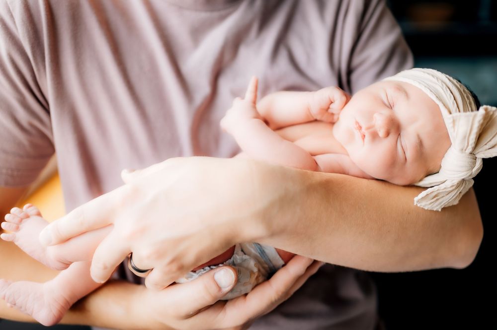 A sleeping newborn wearing a white headband rests peacefully in gentle hands.