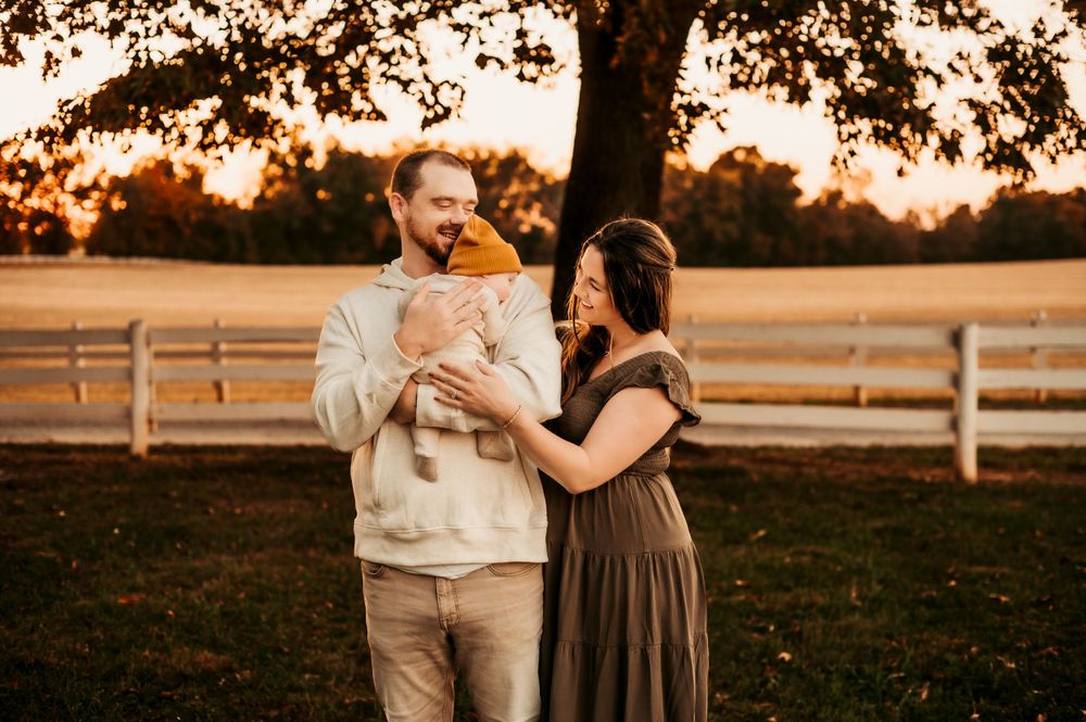 A couple holding their baby while standing in a field at sunset with white fence in background.