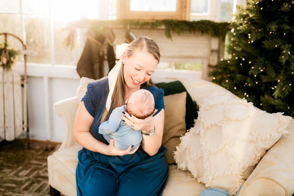A tender moment between parent and baby on a couch next to a Christmas tree in warm sunlit setting.