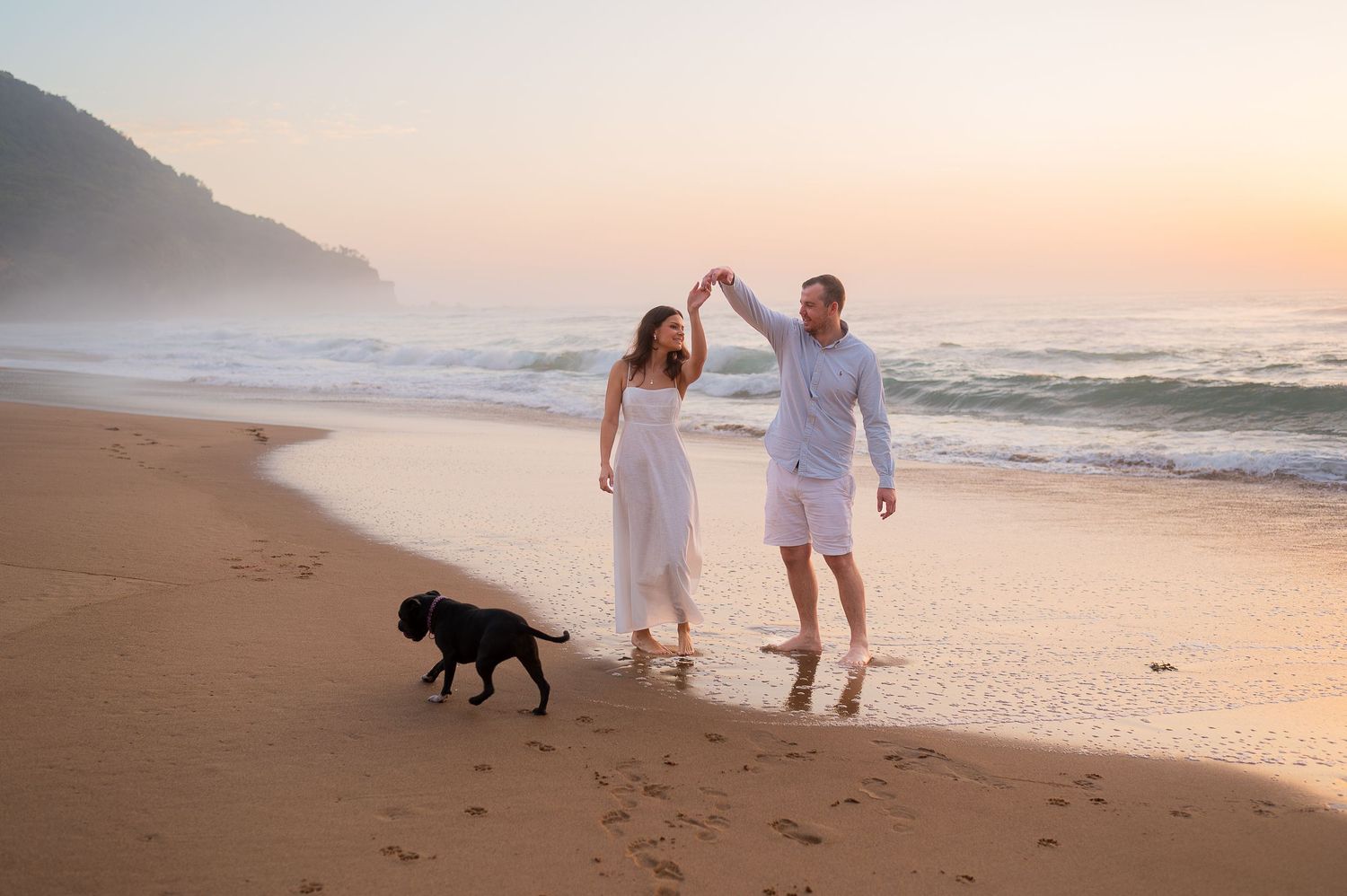 Couple in white dancing on a sandy beach at sunset with a black dog, ocean waves and misty mountains in the background.