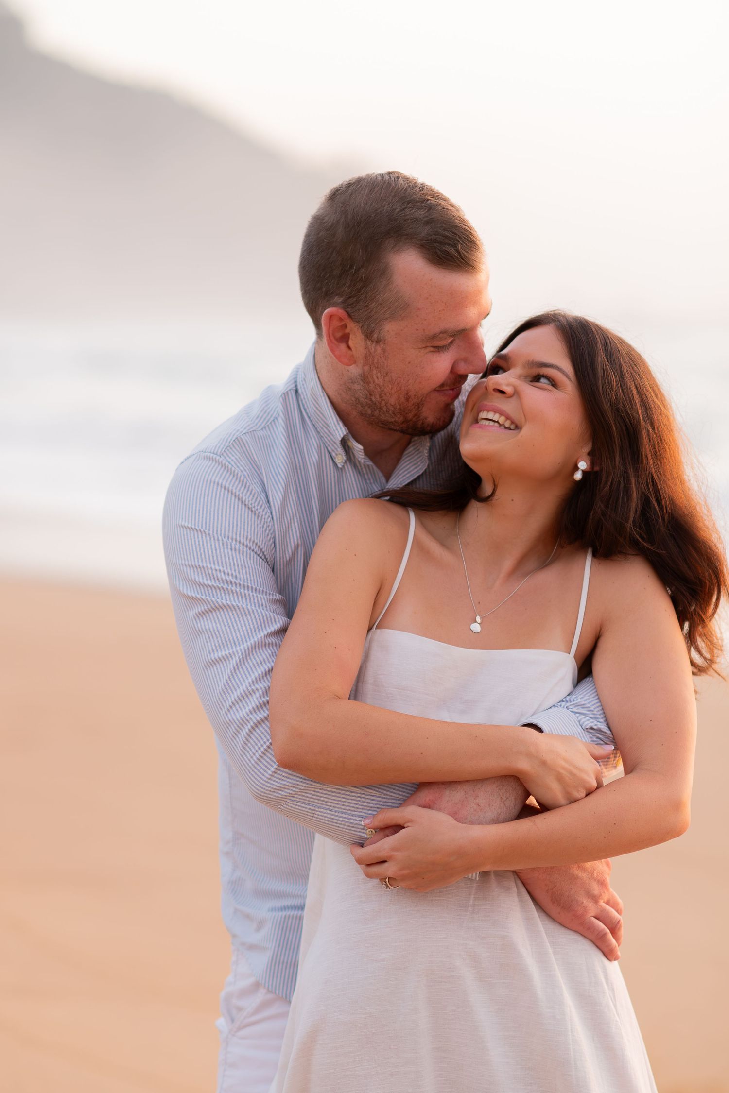 Romantic couple embracing on a beach at sunset, woman smiling as man kisses her cheek.