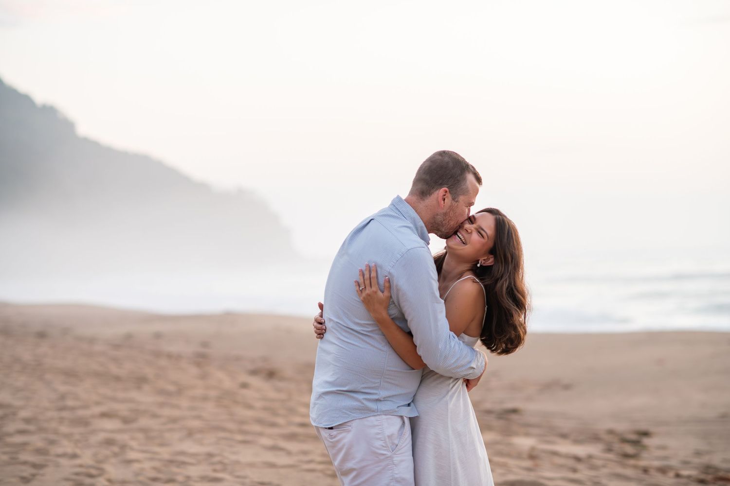 A couple shares a romantic embrace on a sandy beach with misty ocean waves and coastal cliffs in the background.
