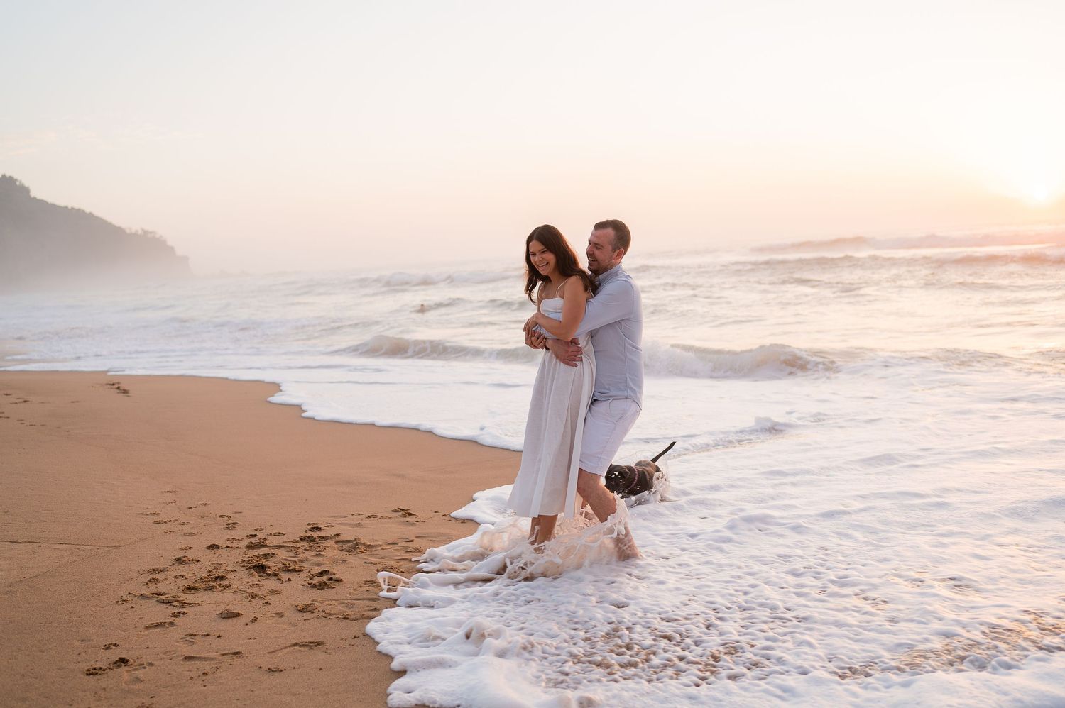 Couple in white embracing on a sandy beach at sunset with a dog playing in the ocean waves.