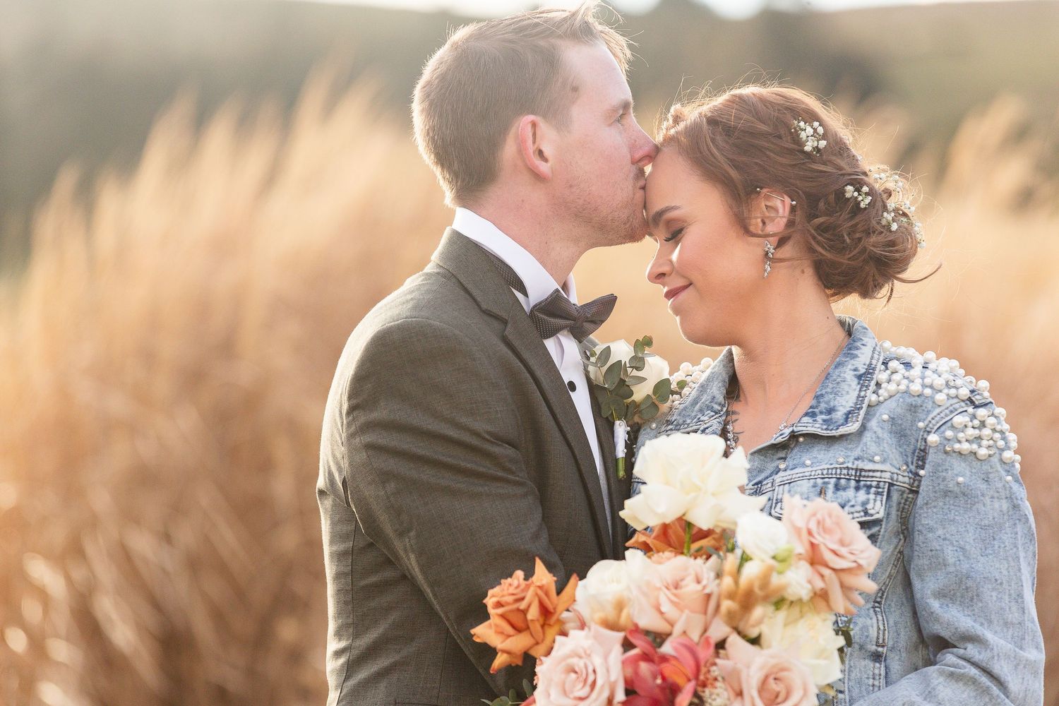 A bride in a denim jacket holds peach and cream flowers while sharing an intimate moment with her groom at sunset.