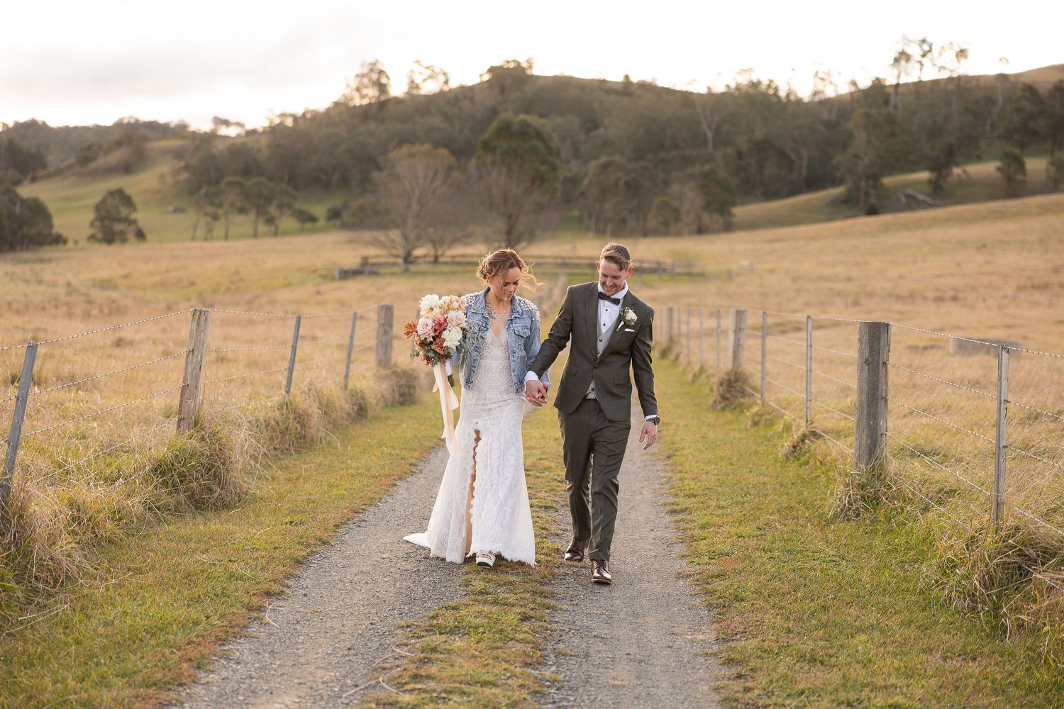 Wedding party walks down a rustic dirt path through golden fields at sunset with mountains in the background.