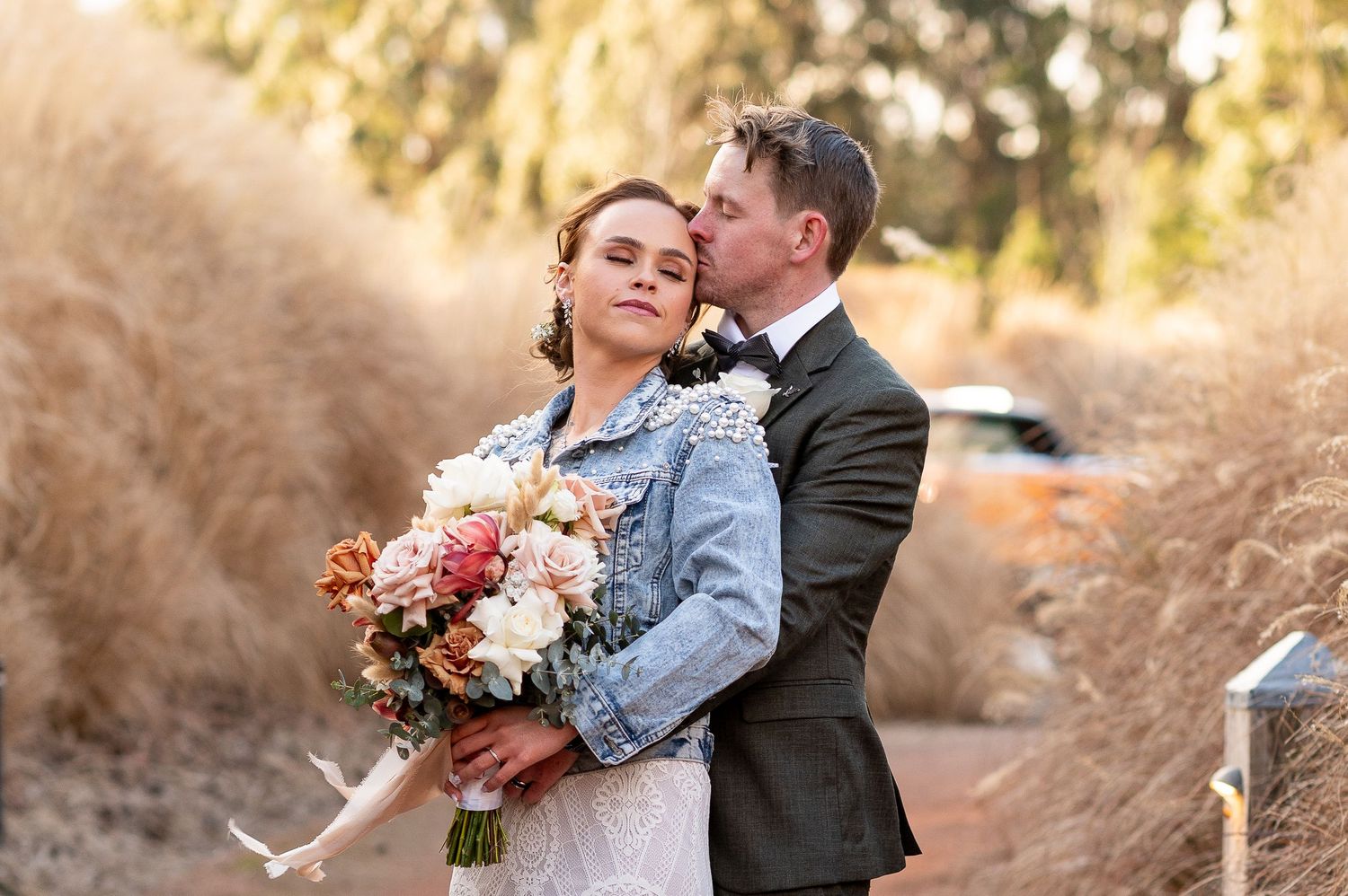 Bride wearing denim jacket holds colorful bouquet while embracing groom in suit during outdoor autumn photoshoot.